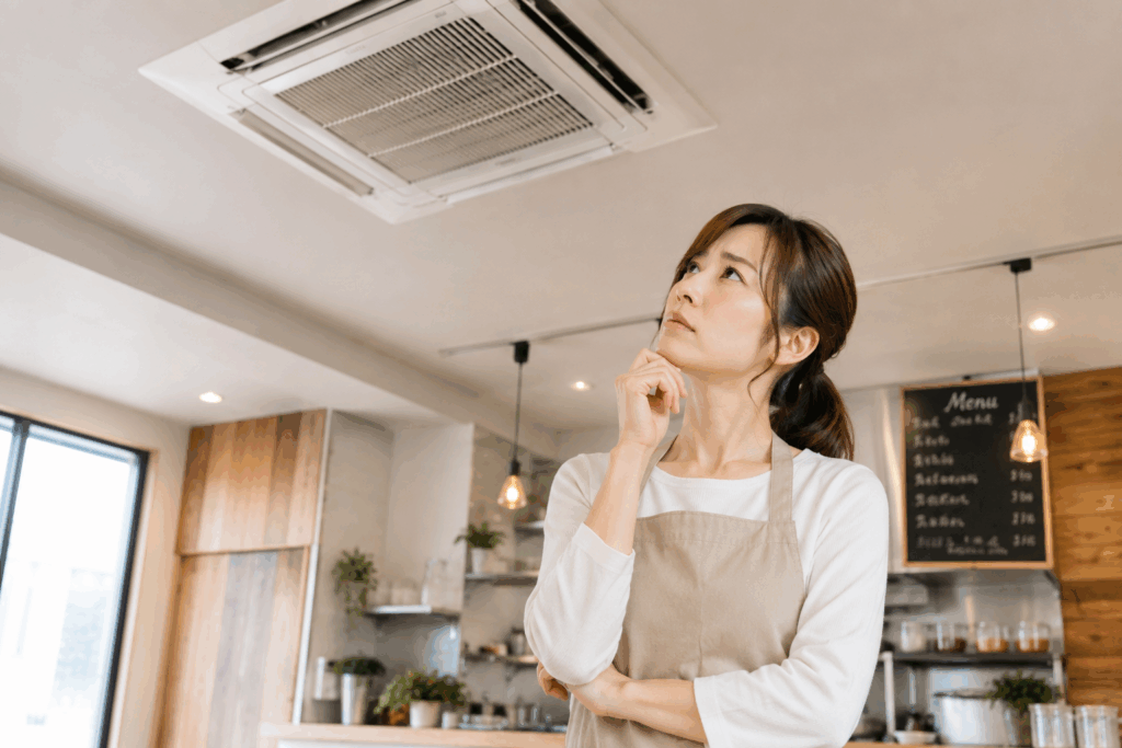 店舗内で40代女性オーナーが天井カセット型の業務用エアコンを見上げながら、冷房の効き具合について考えている様子