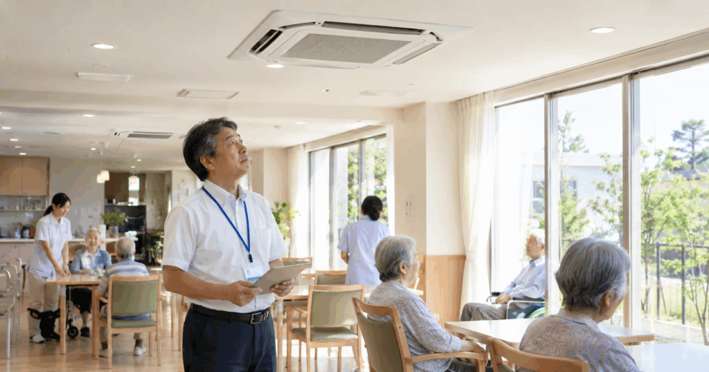介護施設の施設責任者が天井埋込型の業務用エアコンを見上げて室内の空調状態を確認している様子
