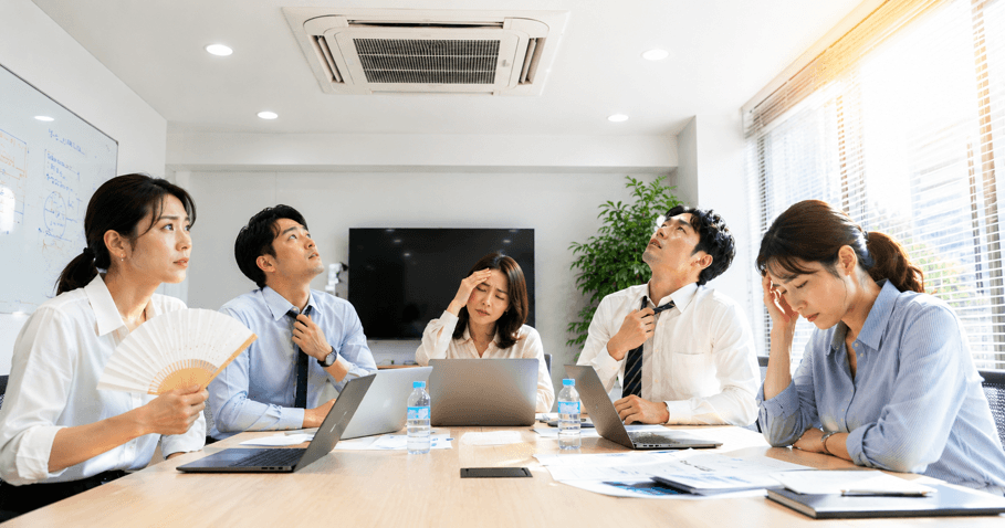会議中に天井カセット型業務用エアコンを見上げながら、暑さでネクタイを緩める男性社員と集中できないオフィススタッフ